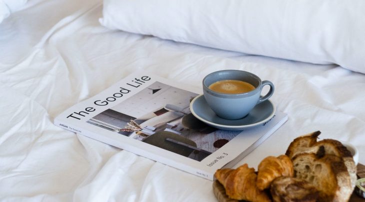 A bed with a wooden tray of bread, croissants, cup of coffee, and magazine