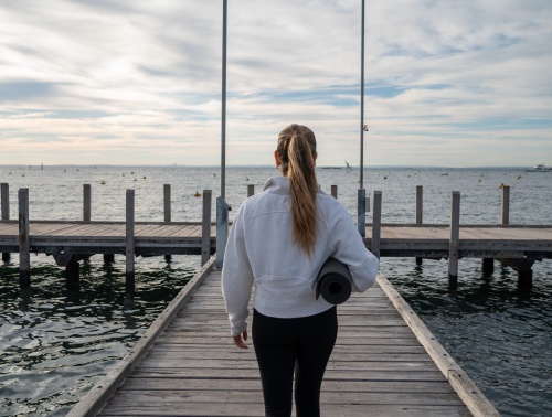 Woman standing on a pier facing the ocean with a yoga mat